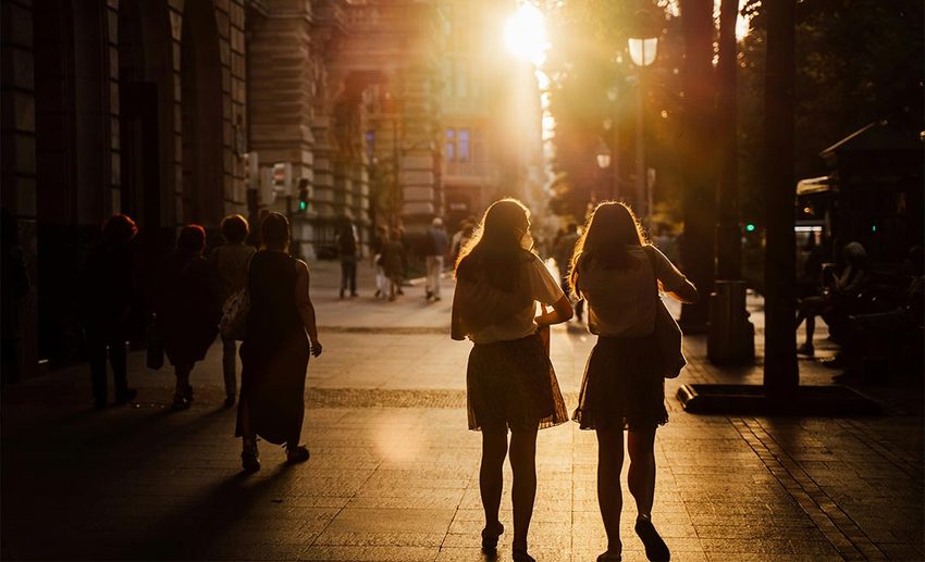 Silhouettes of two women walking together against bright golden sunlight streaming through city streets in Bilbao, Spain.