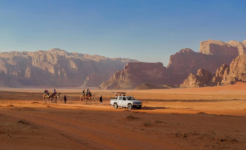 A group of diverse travellers riding in the back of an open-top truck through the red sand dunes of Wadi Rum, Jordan.