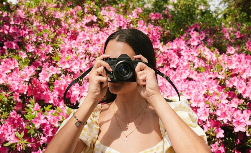 Woman holding a professional camera up to her face to take a photograph in front of vibrant pink flowers.