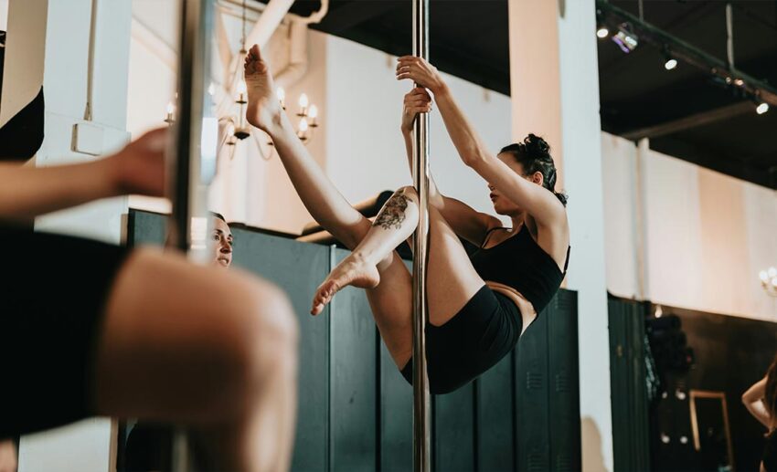 Athletic woman executing an inverted upside-down pose on a dance pole inside a fitness studio.