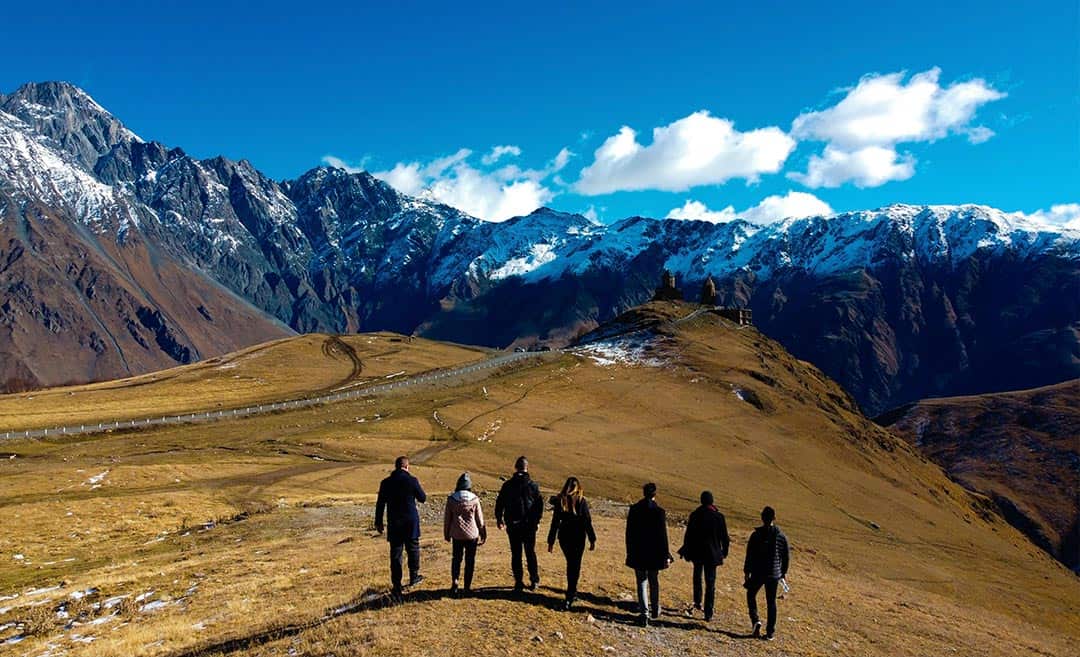Travellers standing in a field with a snow-covered mountain range in the background.