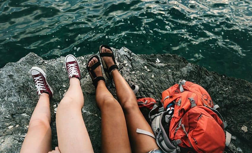 Two backpackers sitting on a high cliff edge overlooking the ocean with their backpacks resting beside them.
