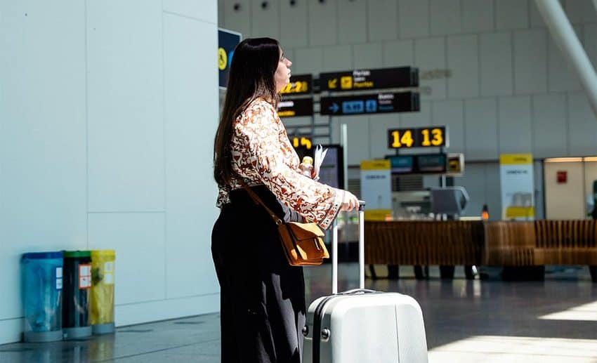 A female traveller with a suitcase looking up at a flight departure board inside an airport terminal.