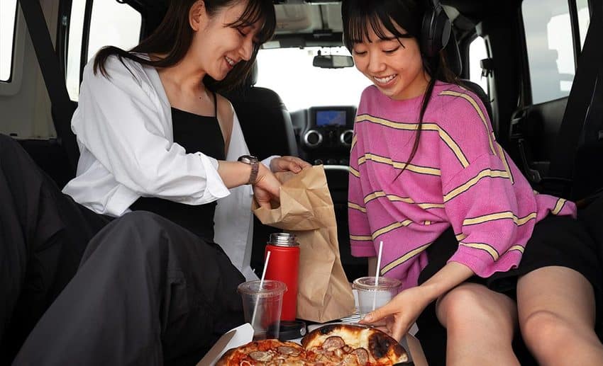 Two women sitting comfortably inside a car on a road trip, eating pizza together during a rainy day.