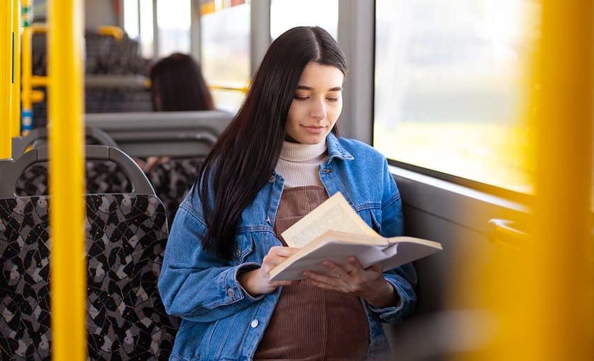 Medium shot of a woman calmly reading a book while seated on a public bus during her travels.