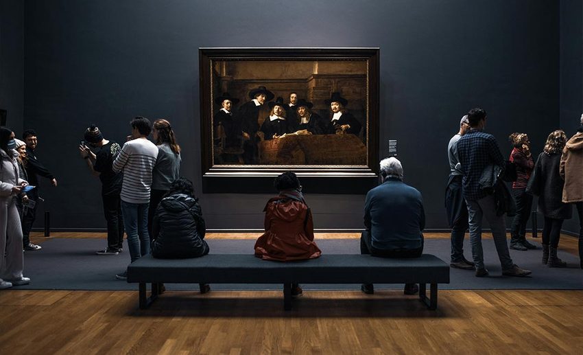 A group of museum visitors, with senior tourists seated on benches, admiring artwork inside the Rijksmuseum in Amsterdam.