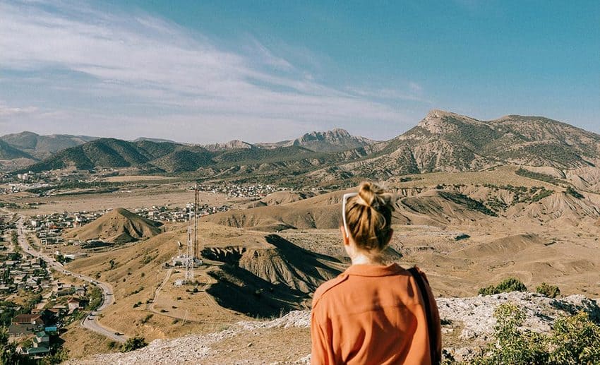 A woman standing at a mountain summit looking out over a wide, sweeping valley below.