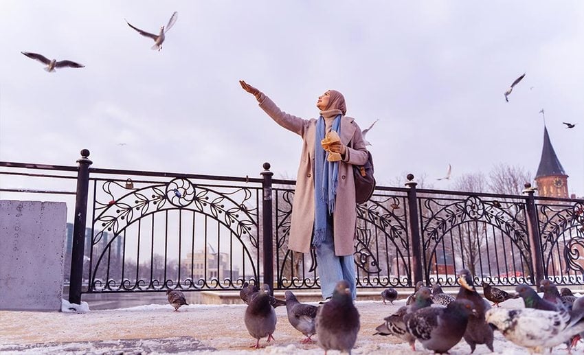 A Muslim woman smiling while feeding pigeons in a vibrant city square during her travels.