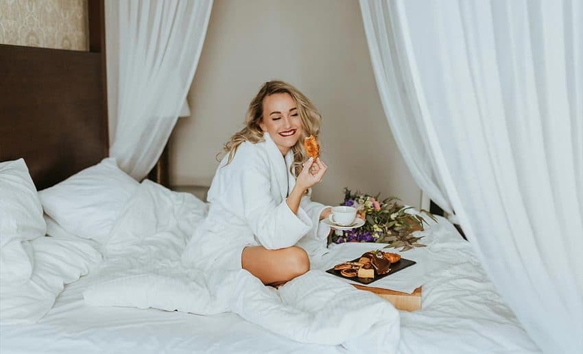 A woman in a white bathrobe sitting comfortably on a plush hotel bed.
