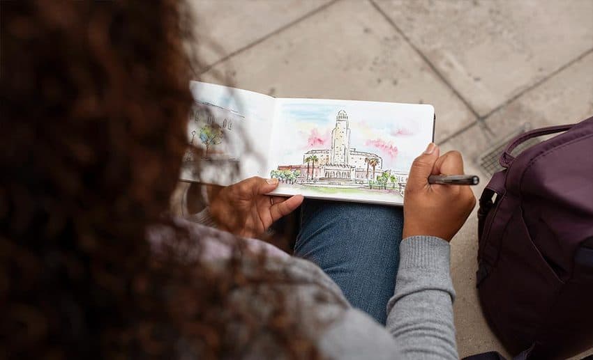 A woman sitting outdoors in an urban setting, engaged in on-the-spot sketching in a notebook.