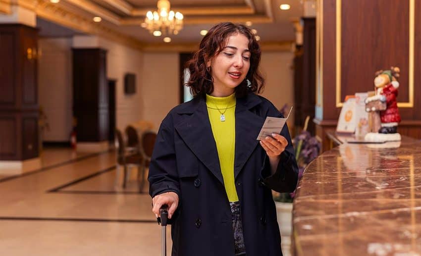 A woman in a dark trench coat checking her keycard folder at a hotel reception desk.