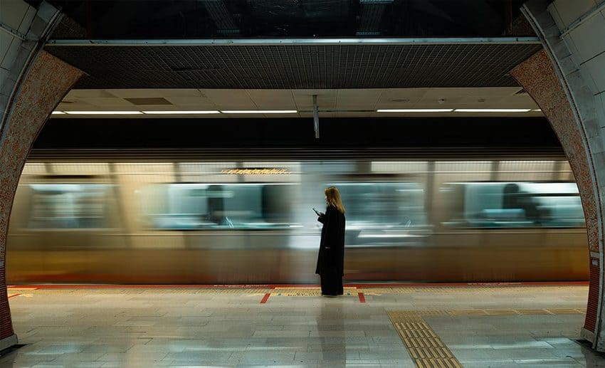A woman standing thoughtfully on a busy train platform with a blurred passenger train passing by.