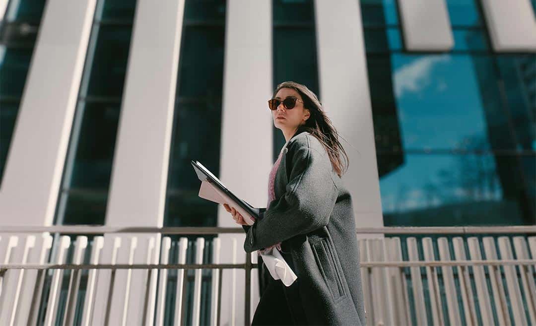 A woman holding documents walking past modern high-rise architecture on a business trip.