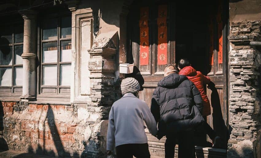A young woman holding hands with her senior parents as they walk through an ornate wooden doorway in Shanghai, China.