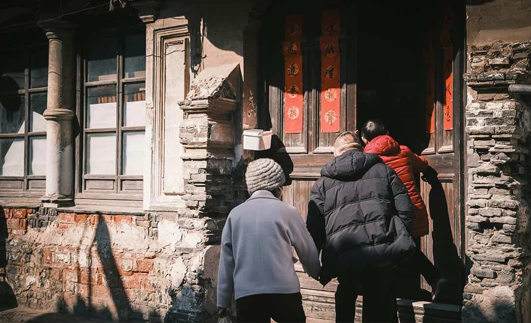 A young woman holding hands with her senior parents as they walk through an ornate wooden doorway in Shanghai, China.